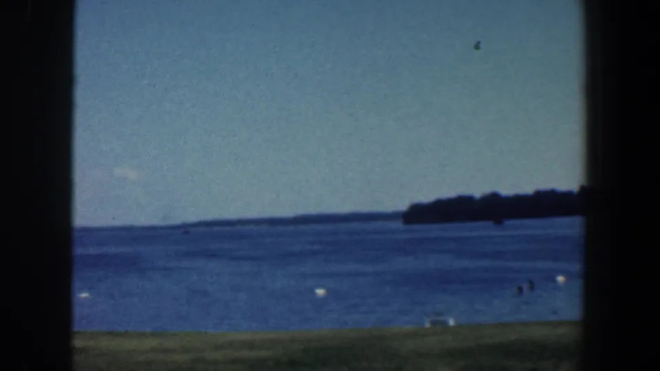 1961 Baltimore-area home-movie frame showing a calm lake, distant wooded shoreline, small people near the water, buoys, and grass.