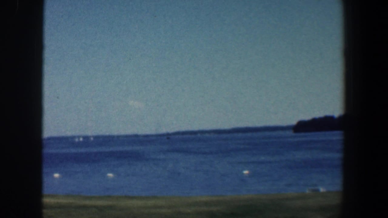 1961 8mm lakeshore view near Baltimore, Maryland, with blue water, white buoys, distant wooded land, and dark frame edges.