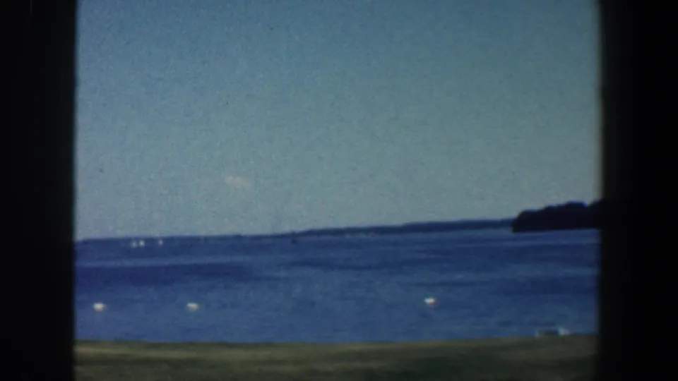 1961 8mm lakeshore view near Baltimore, Maryland, with blue water, white buoys, distant wooded land, and dark frame edges.