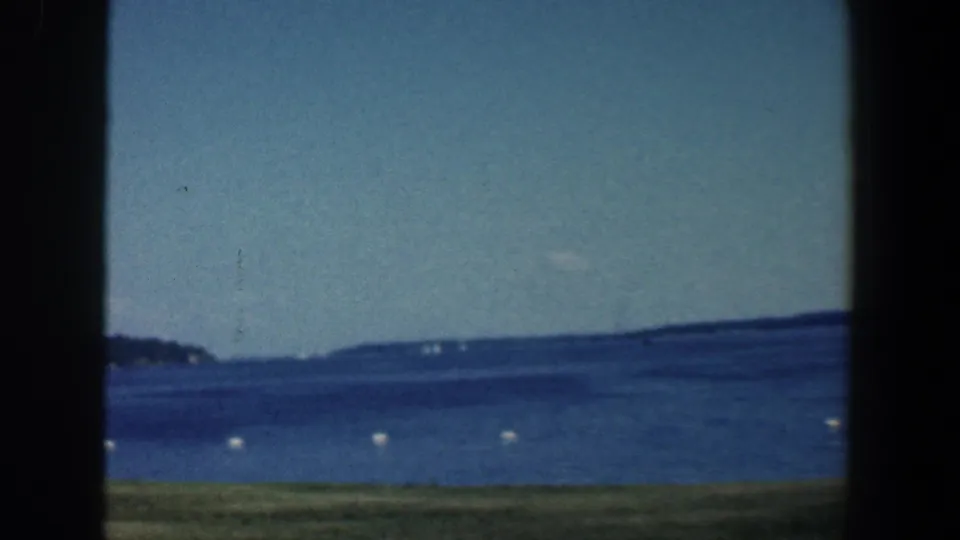 Window-framed 1961 8mm view of a broad blue lake near Baltimore, with buoy markers and low wooded shoreline in the distance.