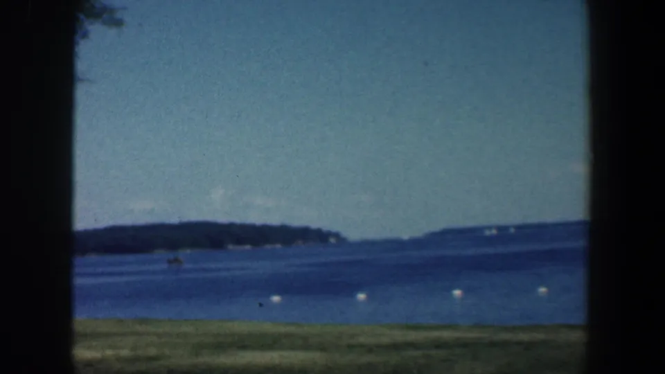 1961 8mm view from a grassy Maryland lakeshore, showing blue water, buoy markers, and a wooded far bank under a clear sky.