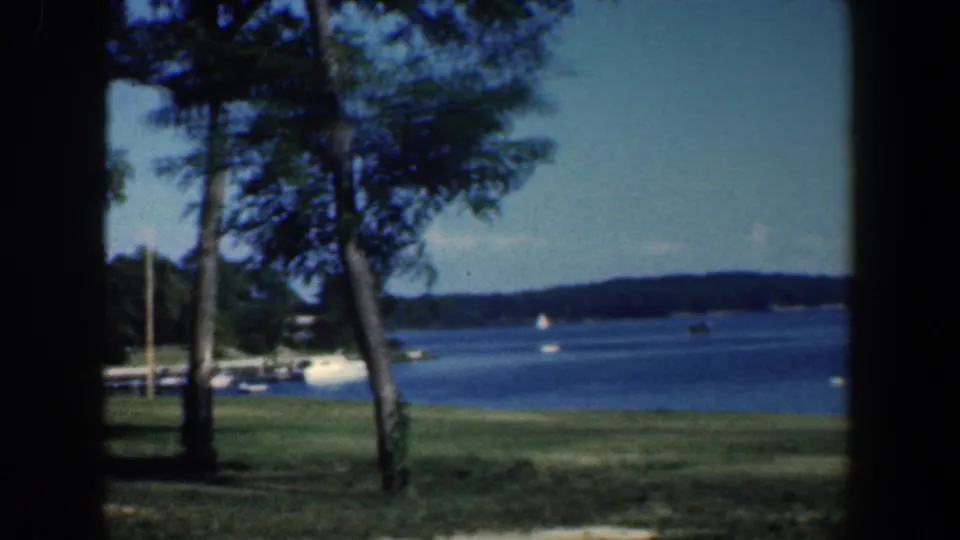 1961 Baltimore-area 8mm frame showing a tree-lined grassy lakeshore, blue water, distant shore, and small boats.