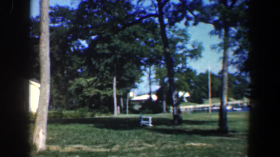 1961 Baltimore-area 8mm frame of a shaded lakeside lawn with trees, dock area, boats, and part of a white building.