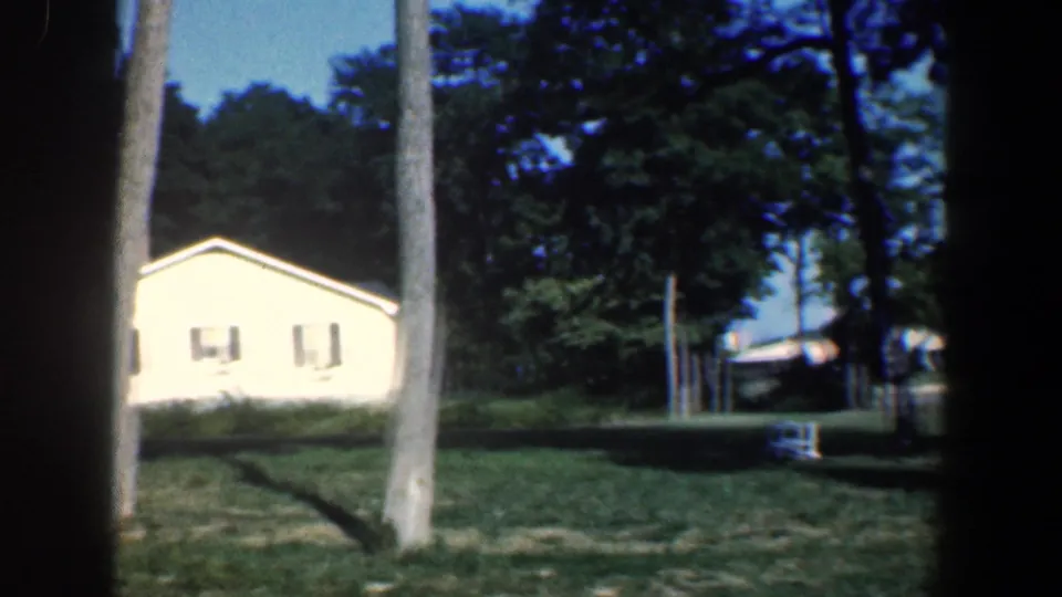 1961 Baltimore-area frame showing a white lakeside house, trees, lawn, dock structures, boats, and glimpses of water.