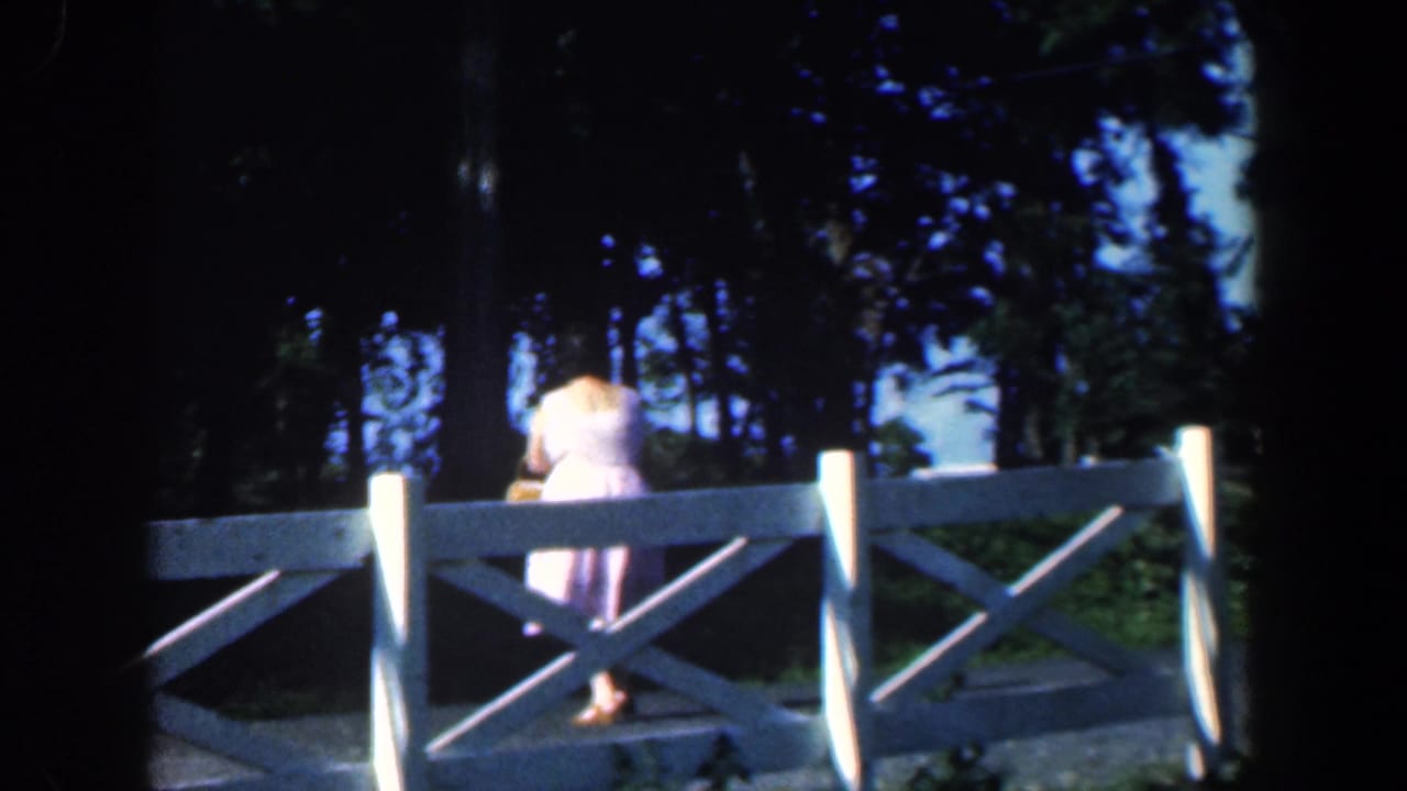 Woman in a light dress walking beside a white wooden fence under trees.