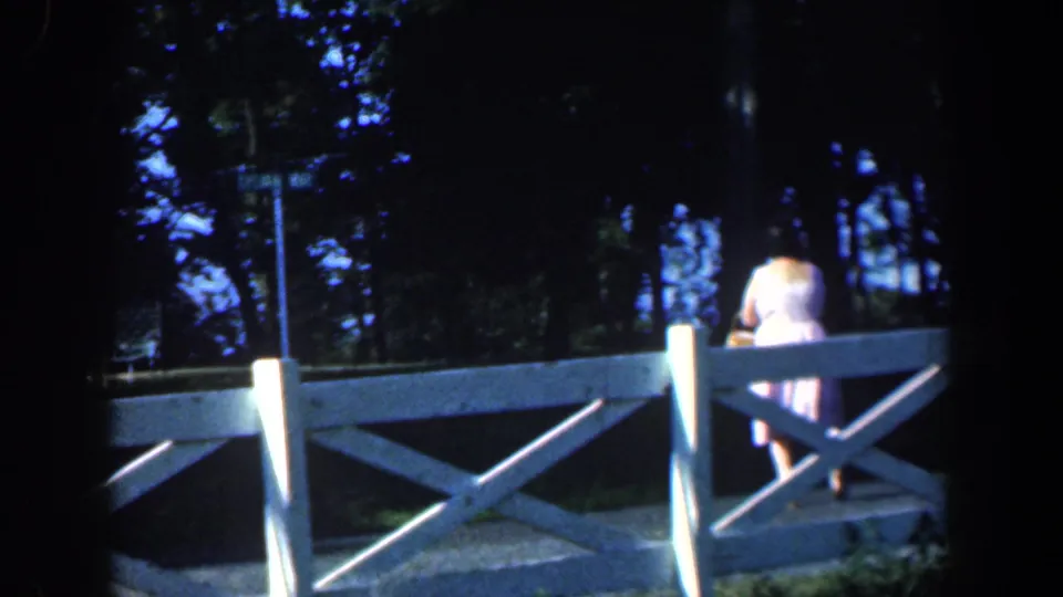 Handheld 8mm view of a woman walking away along a white fence near trees in the Baltimore-area footage.
