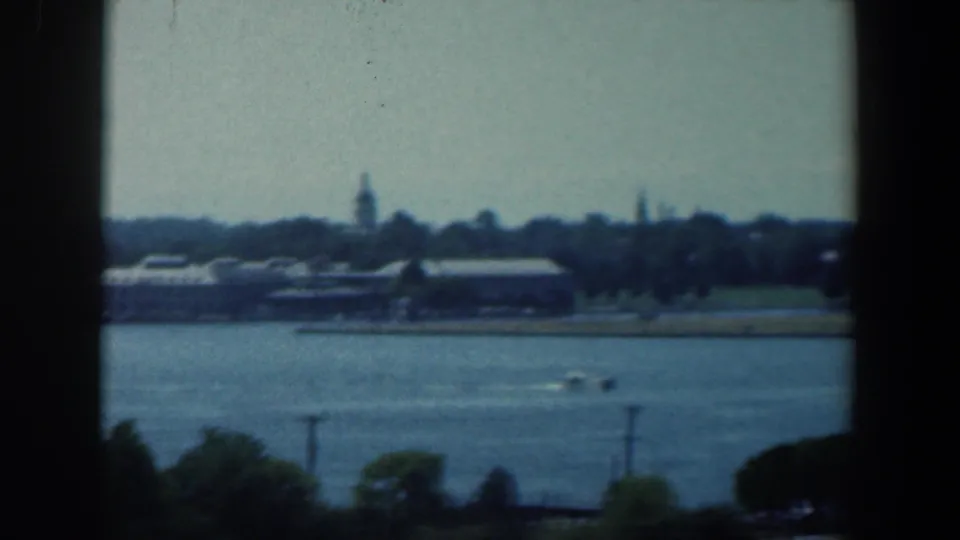 A small speedboat moves across the water in front of the distant Baltimore shoreline, with pale buildings and trees beyond.