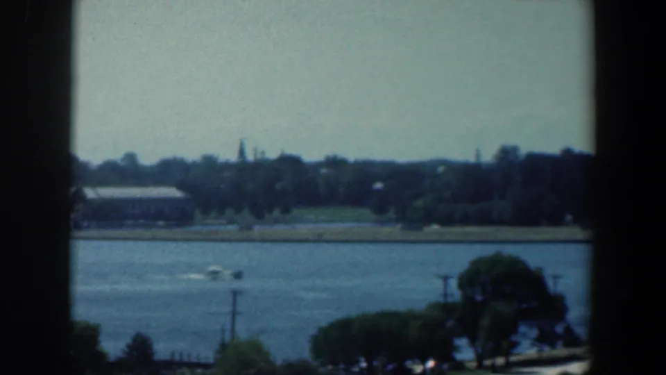 Small speedboat leaves a pale wake across Baltimore harbor water, with trees, low buildings, and dark foreground framing visible.