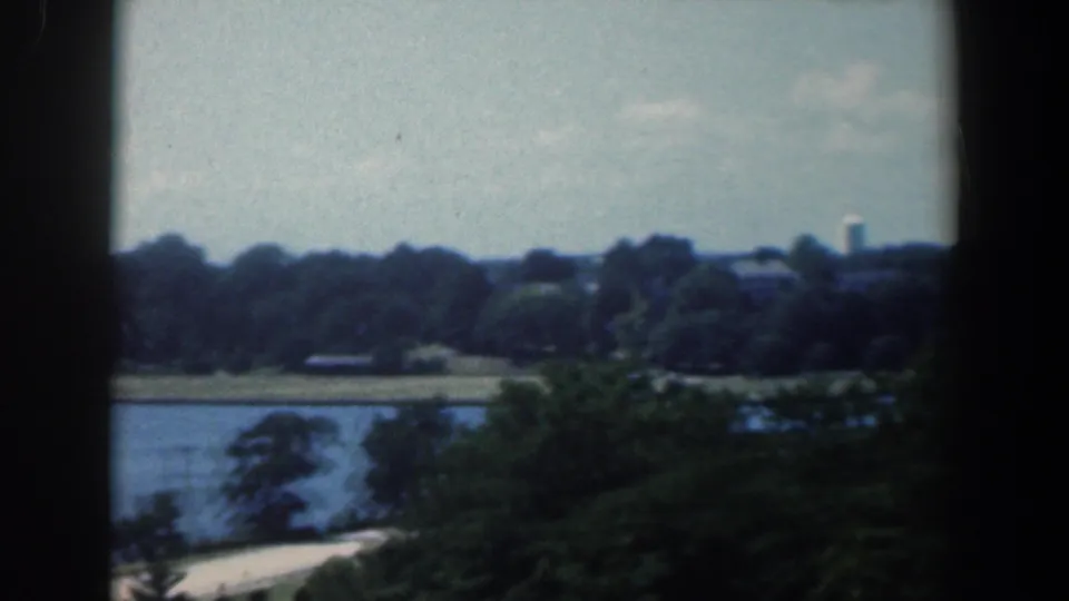 1961 Baltimore 8mm view of a waterfront road, trees, distant buildings, and water, framed by dark vertical edges.