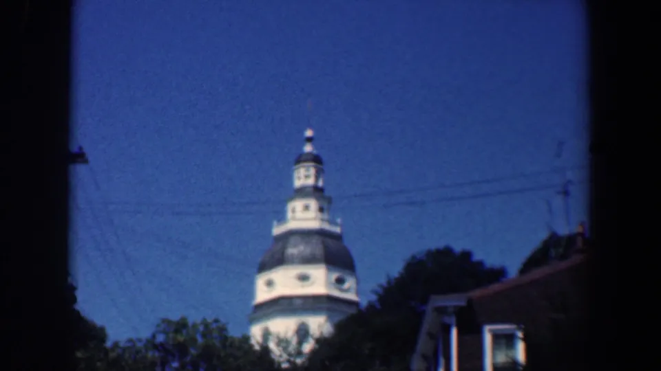 1961 Baltimore frame showing a white church tower with a dark domed roof rising above trees and nearby buildings.