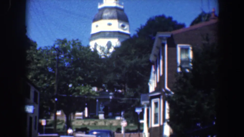 1961 Baltimore home-movie frame showing a domed tower behind trees, house facades, and a partial street edge.