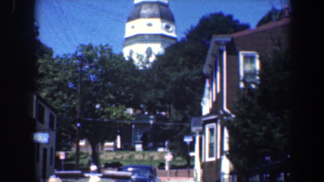 White church steeple above trees, with a brick house and parked car on a Baltimore street.