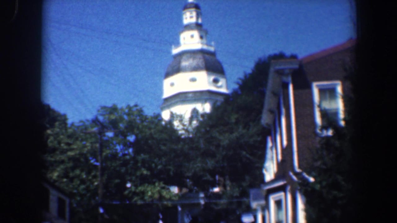 Distant church steeple above trees beside a brick building under blue sky.