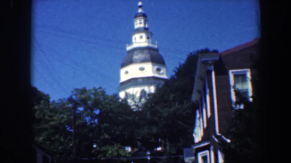 1961 Baltimore home-movie frame with a church dome and steeple above green trees beside a brick residential facade.