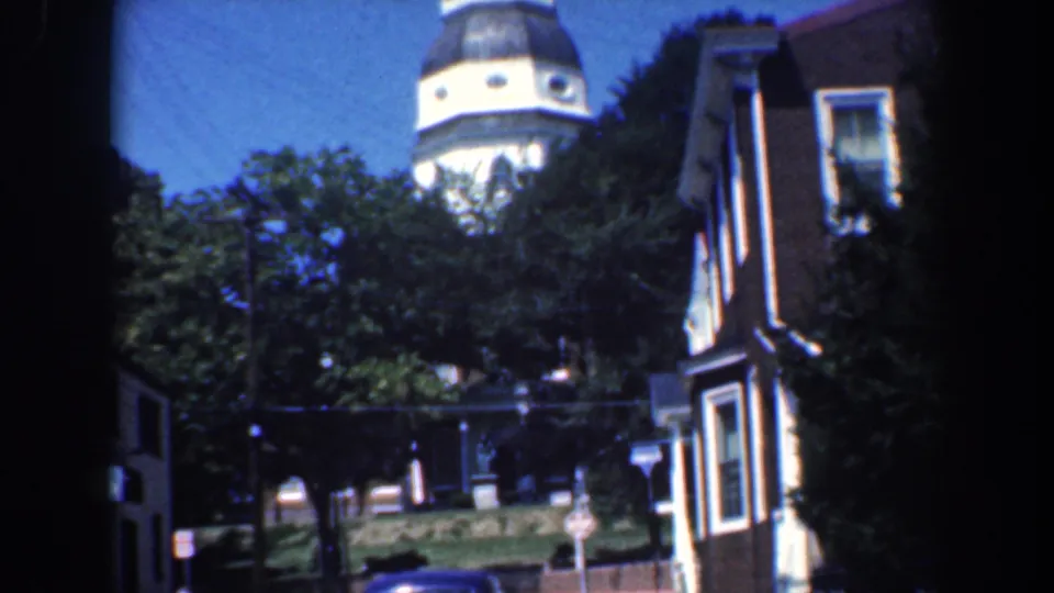 1961 Baltimore street scene showing a distant white church steeple, leafy trees, a brick house, sidewalk area, and a parked car.