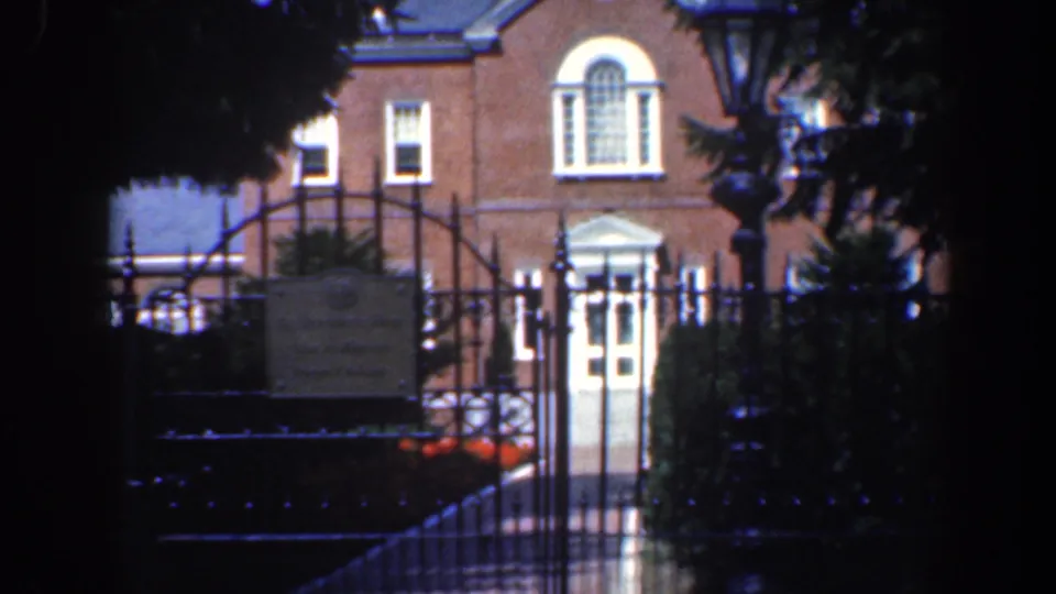 Late in the clip, the gated brick building entrance remains framed by foliage and a foreground lamppost.