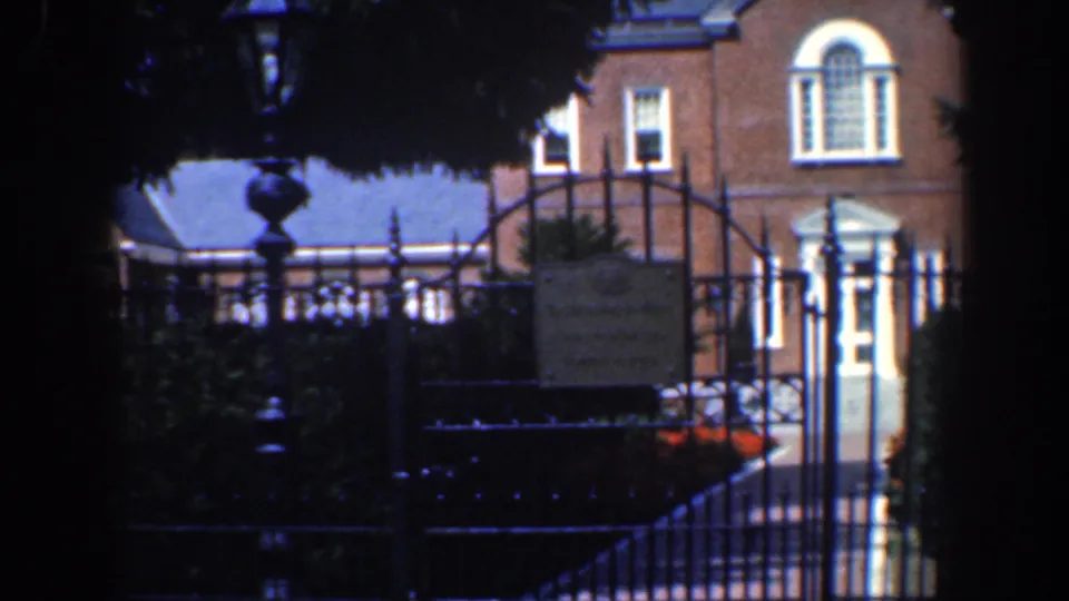 1961 Baltimore home-movie view of a brick house facade, white entrance, iron gate, and shadowed garden foreground.