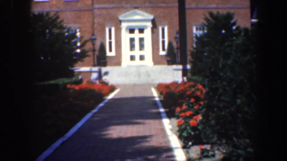 1961 Baltimore 8mm frame with a paved walk, red flowerbeds, shrubs, and a white-trimmed doorway on a brick house.