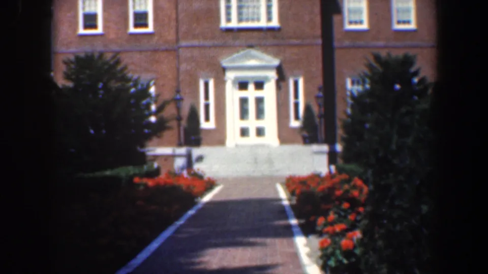 1961 Baltimore exterior frame showing red flowerbeds, manicured shrubs, a paved path, and a white entrance on a brick residence.