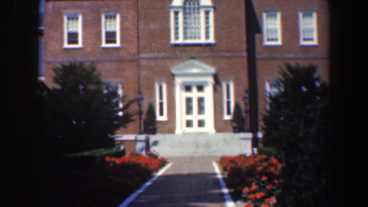 Brick building entrance with white portico, walkway, red flowers, and trees.