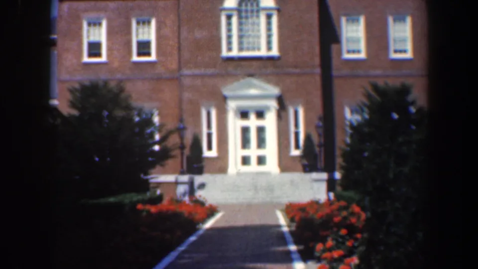 1961 Maryland 8mm frame showing a formal brick building approached by a walkway lined with red flowers and trees.
