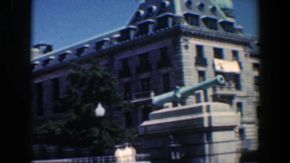 1961 Baltimore home-movie frame with a closer angled view of the cannon monument, stone facade, roofline, and nearby tree shade.