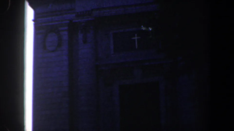 Low-light 8mm frame of a Baltimore church entrance with a visible cross, brick facade, and bright vertical light source.