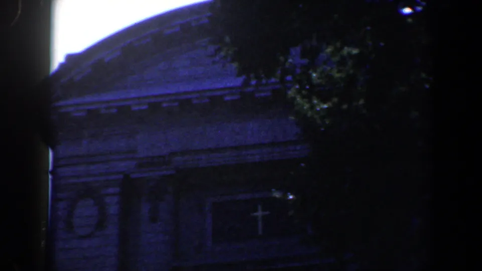 Final 1961 Baltimore 8mm frame of the shaded church front, with the pediment, cross motif, and tree leaves visible against bright sky.