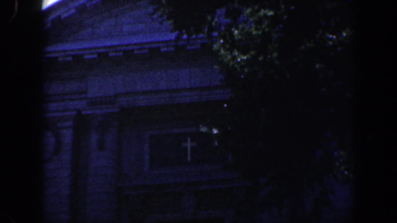 Dark view of a masonry building partly hidden by trees, with a cross-shaped emblem visible.