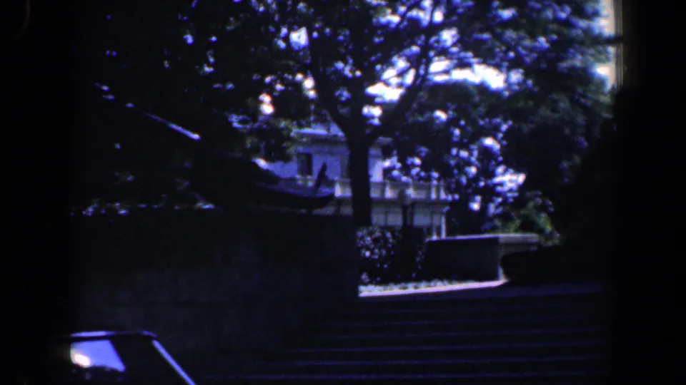 1961 Baltimore frame showing a partial car roof in the foreground with shaded stone steps, trees, and a building beyond.