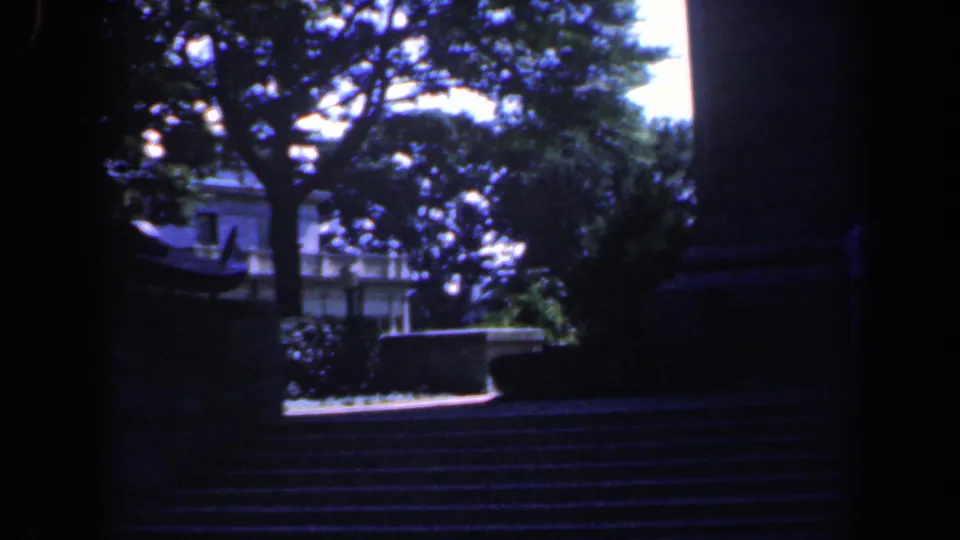 1961 Baltimore 8mm frame with a large tree, low wall, stone steps, and sunlit building details in the distance.