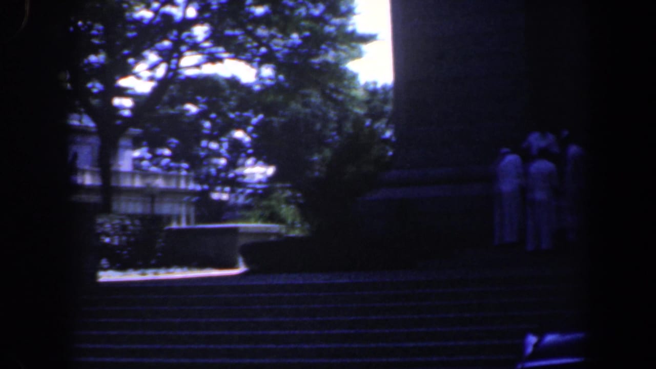 1961 Baltimore home-movie frame showing people in white shirts gathered on stone steps near a large shaded monument or building base.