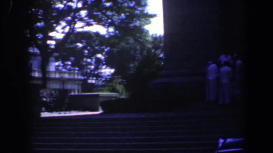 1961 Baltimore home-movie frame showing people in white shirts gathered on stone steps near a large shaded monument or building base.