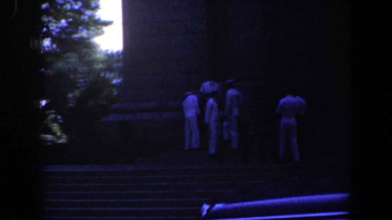 Several people in light clothing stand on steps beside a large stone structure.