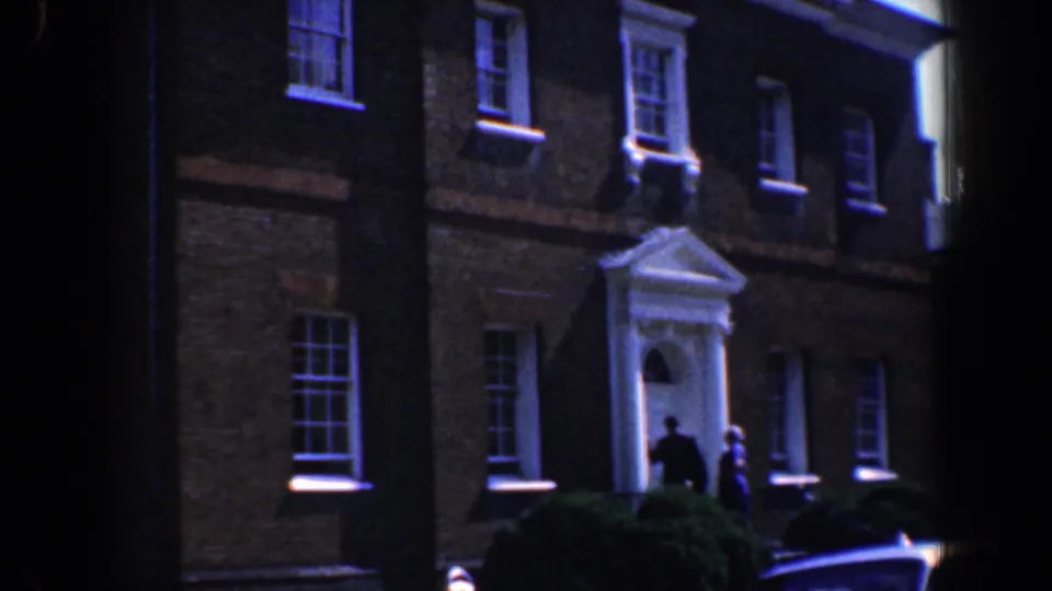 1961 Baltimore frame showing a brick or masonry building with sash windows, a white doorway, shrubbery, and two people near the entrance.