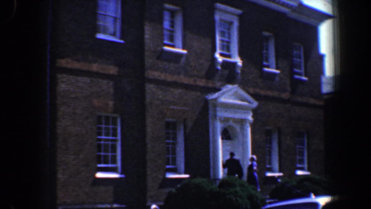 Brick residential building with white portico, two people near the doorway, shrubs, and a parked car edge.