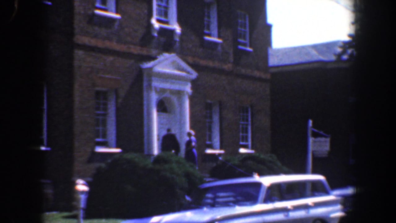 Brick building facade with a white entrance, two people, shrubs, an American flag, and a light station wagon.