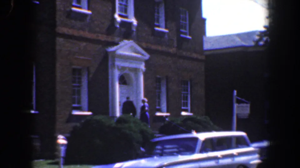 1961 Baltimore residential scene with people near a white entrance, an American flag, shrubs, and a light-colored station wagon at the curb.