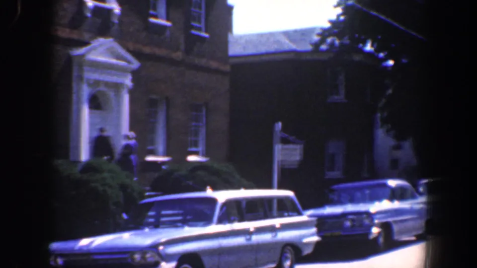 1961 Baltimore street view with a brick building, two figures near the portico, an American flag, and classic cars parked along the curb.