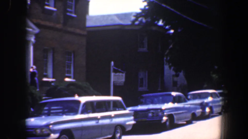1961 Baltimore residential curb with brick building, American flag, shrubs, and several mid-century American cars in bright sun.