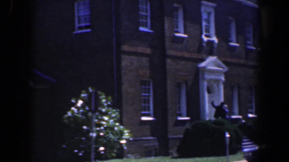 1961 Baltimore exterior with a white portico on a brick building, shrubs, windows, and a person near the entrance steps.