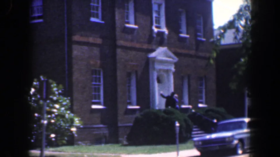 1961 Baltimore street view with brick buildings, shrubs, a sidewalk, and a parked mid-century car in soft 8mm color.