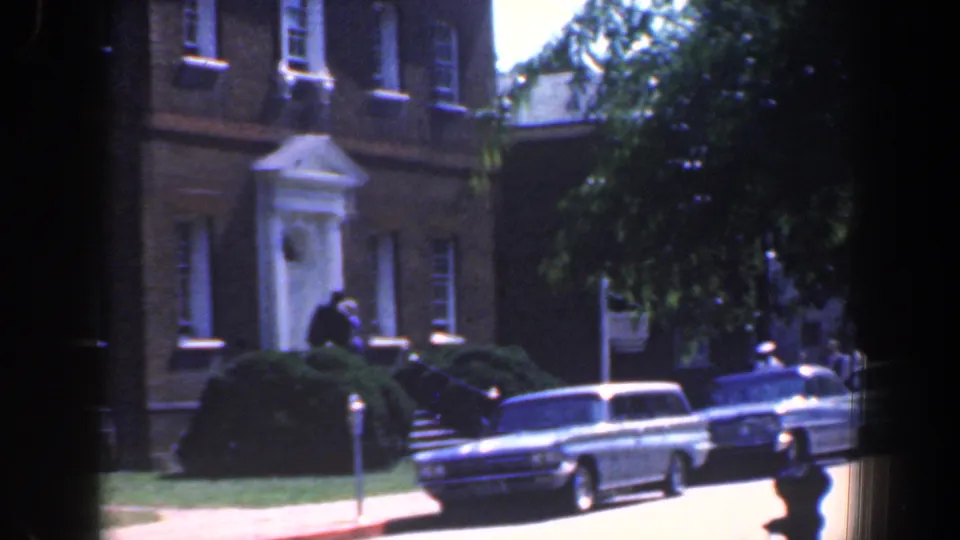 1961 Baltimore street scene with brick buildings, a parked station wagon, shrubs, and a pedestrian near the curb.