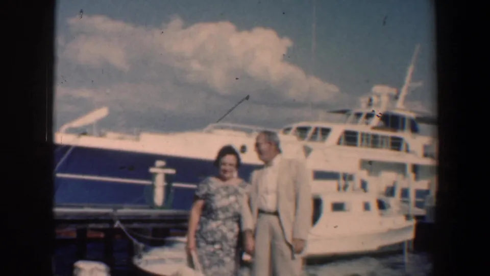 In 1961 Baltimore, an older couple poses on a dock beside a large white yacht, with water and clouds behind them.