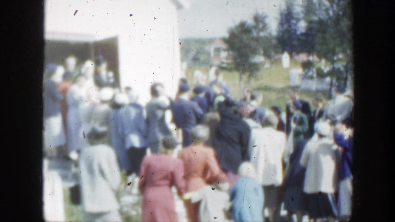 Wedding guests gather outside a light-colored building near a bride and groom.