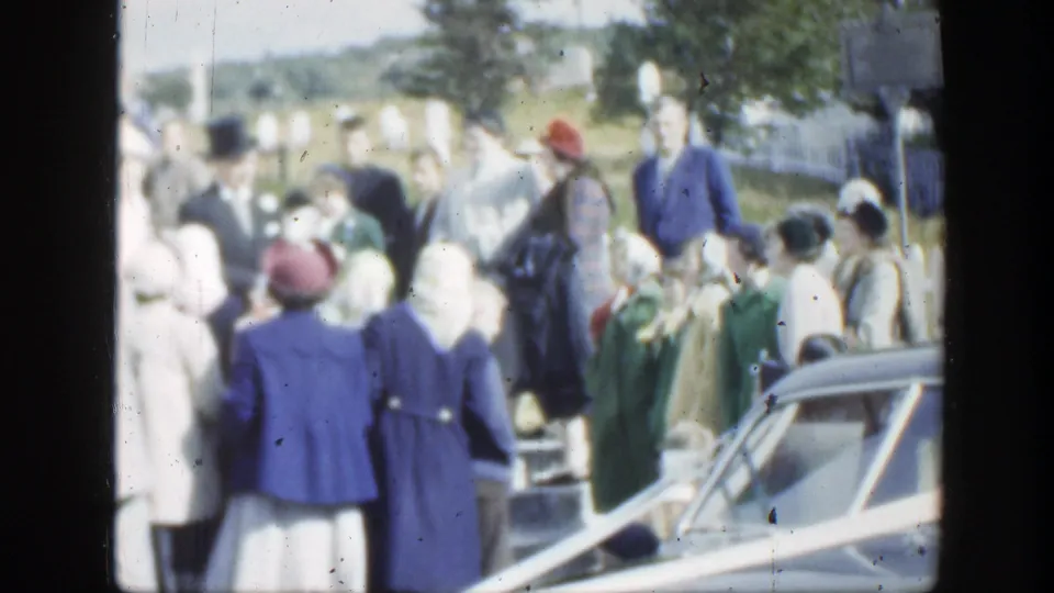1950 Montreal wedding guests in formal clothing gather near a car and flowers, facing the activity around the building.