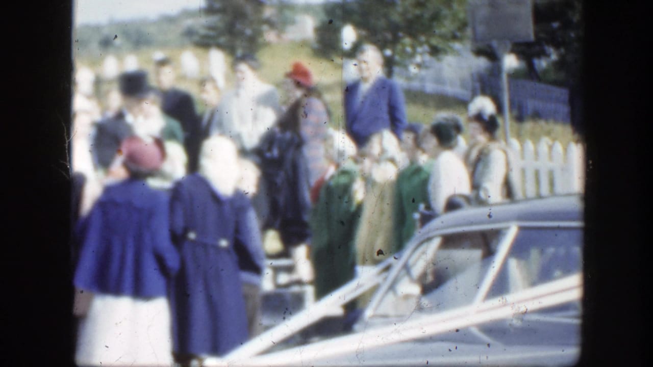 1950 Montreal wedding guests in formal dress gather beside a ribboned vintage car near a white picket fence.