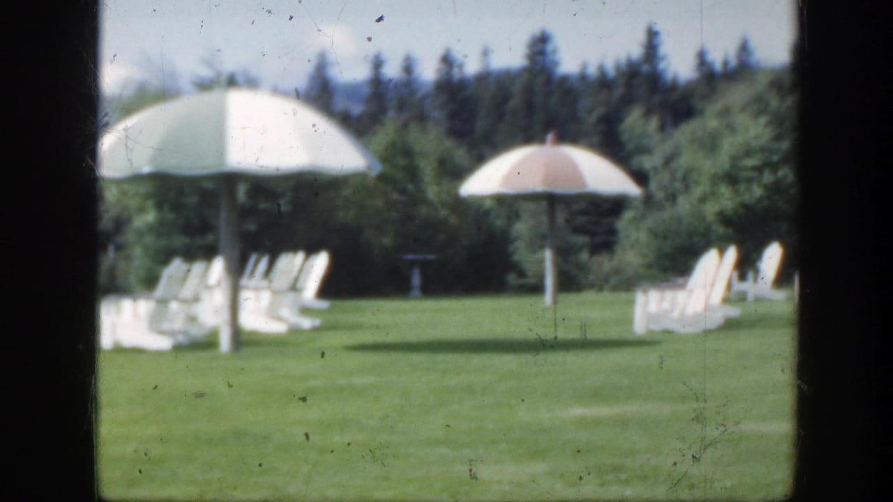 1950 Montreal 8mm frame showing a green lawn with white chairs, parasols, and evergreen trees beyond a dark foreground frame.