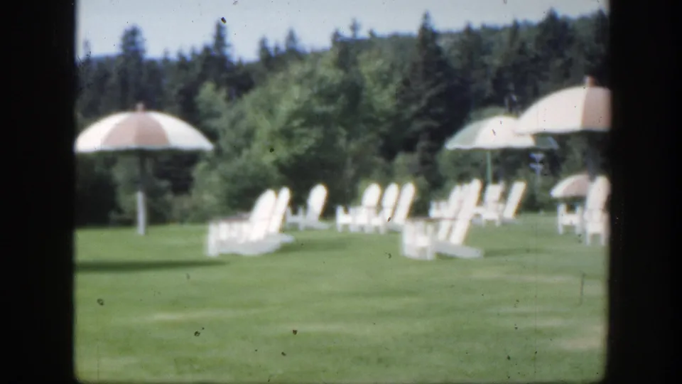 Soft 1950 Montreal home-movie frame with parasols and white chairs scattered across a landscaped lawn.