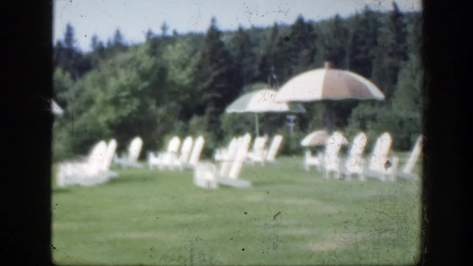 1950 Montreal 8mm frame of empty white chairs and parasols on a green lawn with dense trees behind.